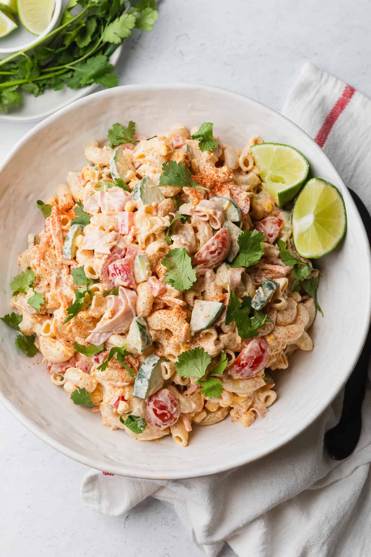 Mexican macaroni salad recipe in a white bowl with lime wedges and cilantro leaves.