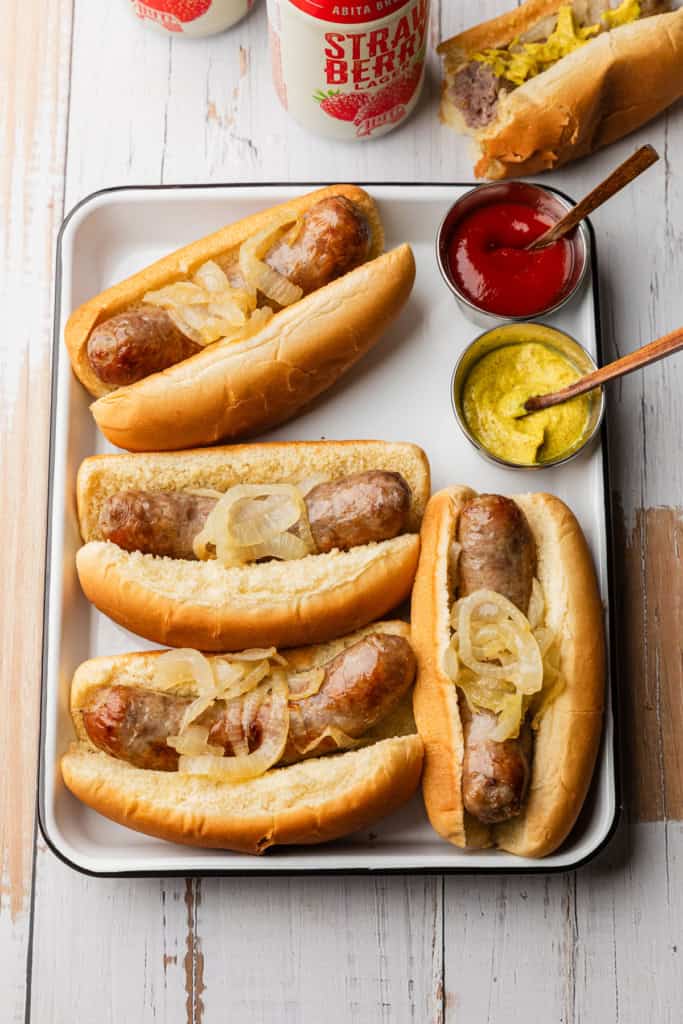 A tray of bratwurst in buns with sliced onions and condiments on the side.