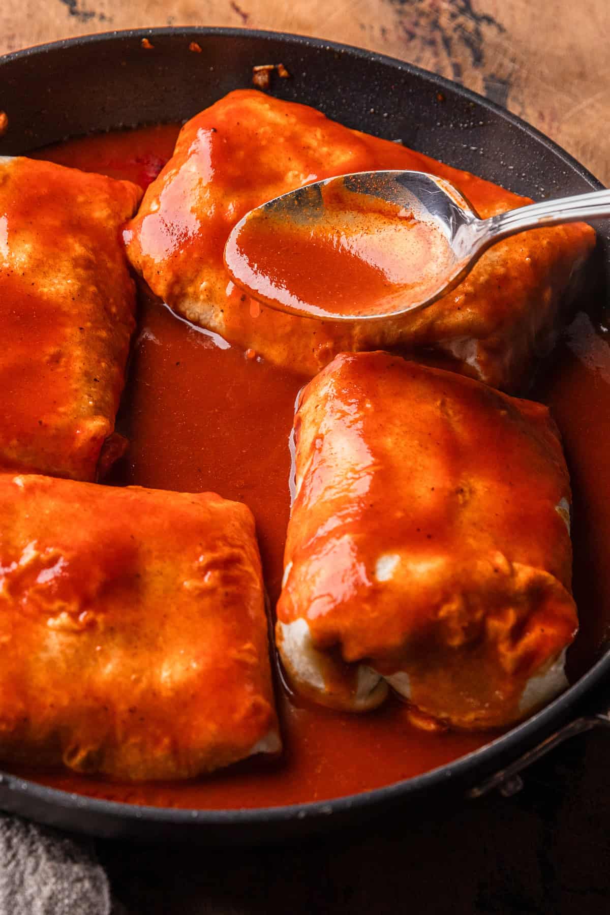 Pouring red enchilada sauce over burritos in a skillet.